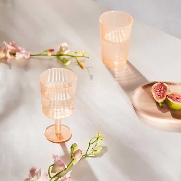 Zwei rosafarbene Weingläser aus Plastik, eine Erdbeere auf einem Holzteller, umgeben von Blumen auf einem Marmortisch.