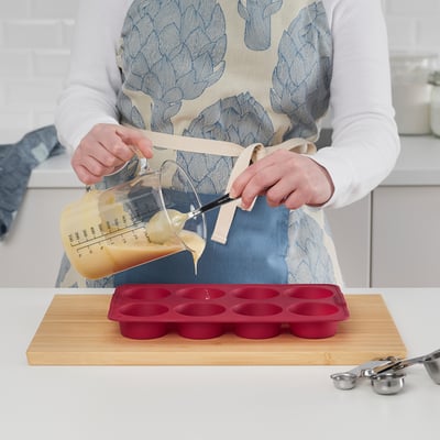 Eine Person gießt Teig in eine rote Silikon-Muffinform, die auf einem Holzbrett steht, und demonstriert so seine Verwendung zum Backen von Muffins oder Desserts.