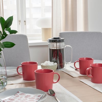 Vier rote Tassen neben einer Kaffeepresse auf einem Esstisch mit Tellern, Servietten und einem Löffel.