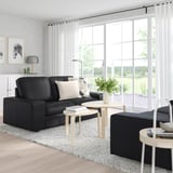 Modernes Wohnzimmer mit großem schwarzem KIVIK-Sofa, Couchtischen aus Holz, weißen Vorhängen und Blick auf grüne Felder durch große Fenster.