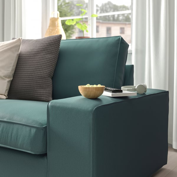 Grünes KIVIK-Sofa mit dunkelgrauen Kissen, Schüssel, Fernbedienung und Kopfhörern an der Armlehne in der Nähe des Fensters.