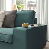 Grünes KIVIK-Sofa mit dunkelgrauen Kissen, Schüssel, Fernbedienung und Kopfhörern an der Armlehne in der Nähe des Fensters.