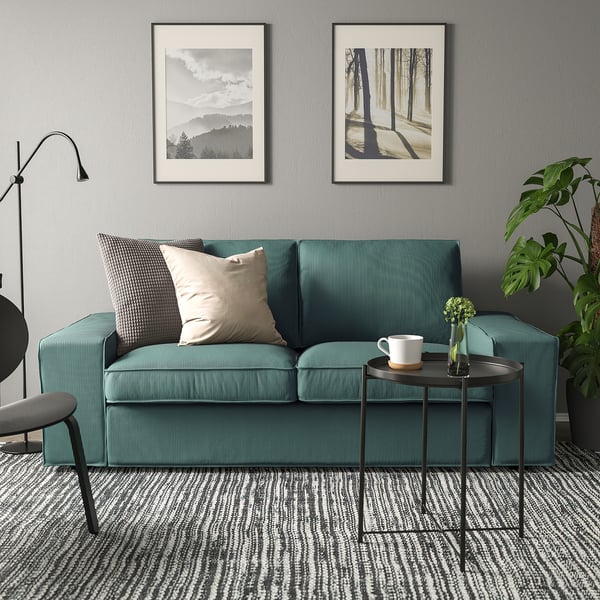 Gemütliche Wohnzimmerszene mit KIVIK-Sofa in Grün, Plüschkissen, kleinem Metalltisch, Pflanzen, Becher und Kunstdrucken an grauen Wänden.