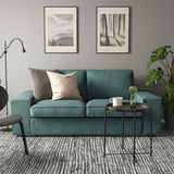 Gemütliche Wohnzimmerszene mit KIVIK-Sofa in Grün, Plüschkissen, kleinem Metalltisch, Pflanzen, Becher und Kunstdrucken an grauen Wänden.