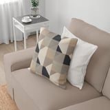 Beigefarbenes KIVIK-Sofa mit buntem Kissen, Beistelltisch mit Kaffeetasse, Pflanze und weißen Vorhängen.
