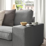 Ein graues KIVIK-Sofa mit einem Kissen und einer Schale auf der Armlehne, in der Nähe eines Fensters mit weißen Vorhängen.