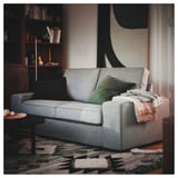 Gemütliches graues KIVIK-Sofa mit grünem Kissen, weicher Decke und abstraktem Teppich in der Nähe eines Bücherregals.
