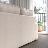 Ein beiges KIVIK-Sofa mit niedrigen Armlehnen und Taschenfedern, das in der Nähe eines Fensters mit Pflanzen gezeigt wird und Komfort und Unterstützung zum Entspannen bietet.