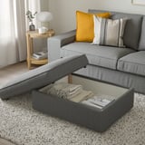 Modernes Wohnzimmer: graues Sofa mit gelbem Kissen, Beistelltisch aus Holz mit Lampe und Büchern. Ottomanische Öffnung mit Decke und Zeitschriften im Inneren.