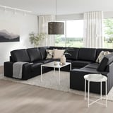 Modernes Wohnzimmer mit L-förmigem schwarzem KIVIK-Ledersofa, großen Fenstern, weißem Dekor, gemütlichen Kissen und Blick auf die Natur.