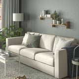 Modernes Zimmer mit hellgrauem KLAVRESTRÖM-Sofa, weißen Kissen, Holzregalen mit Pflanzen und Vasen, grauen Vorhängen, kleinem Tisch, Lampe und Teppich.