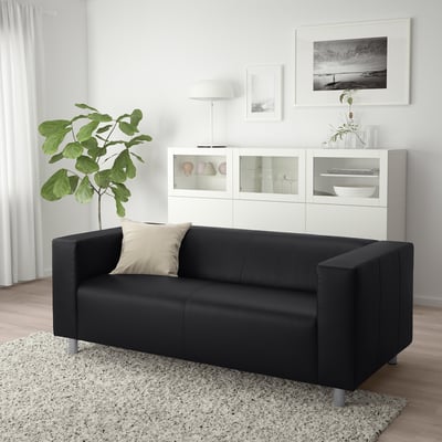 Modernes Wohnzimmer mit schwarzem KLIPPAN-Sofa, beigem Kissen, weißem Schrank und großer Pflanze.