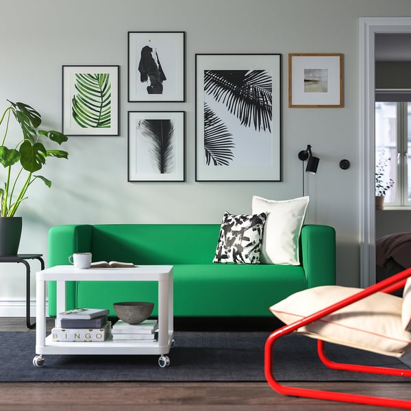 Helles Wohnzimmer mit grünem KLIPPAN-Sofa, trendigen Drucken, weißem Kartell-Tisch und rotem Stuhl.