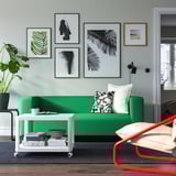 Helles Wohnzimmer mit grünem KLIPPAN-Sofa, trendigen Drucken, weißem Kartell-Tisch und rotem Stuhl.