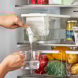 Person, die den durchsichtigen Behälter mit Wasser aus dem Kühlschrankspender füllt, umgeben von Gemüse und Gewürzen.