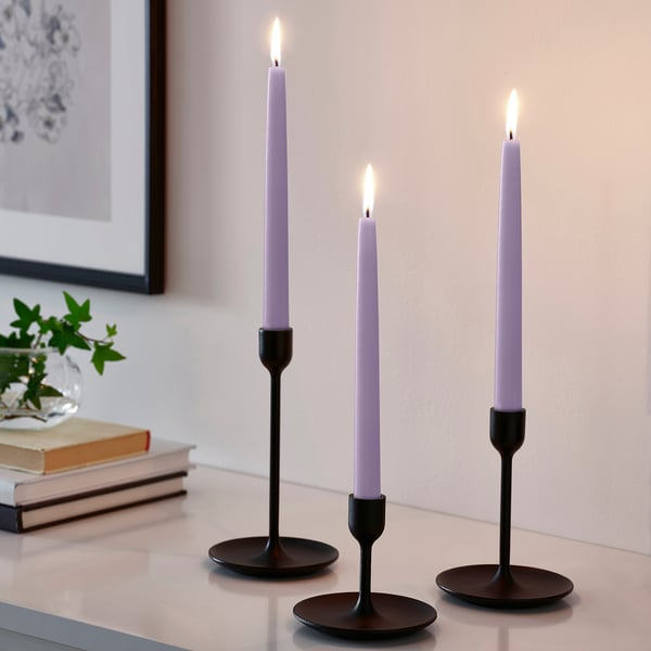 Drei schwarze Kerzenständer mit hohen lila Kerzen und warmem Licht, neben einer Pflanze und Büchern.