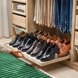 Organisierter Kleiderschrank mit ausziehbarer Schuhablage für schwarze und braune Anzugschuhe. Helles Holzgestell und grün gestreifter Teppich.