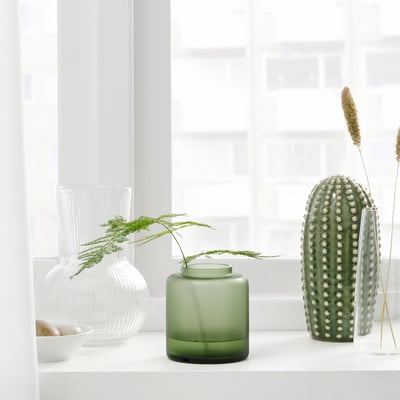 Vier einzigartige Vasen auf einer Fensterbank, die Blumen und Grün in einer minimalistischen Umgebung präsentieren. Zwei klare, eine mattgrüne und eine strukturierte grüne Vase.