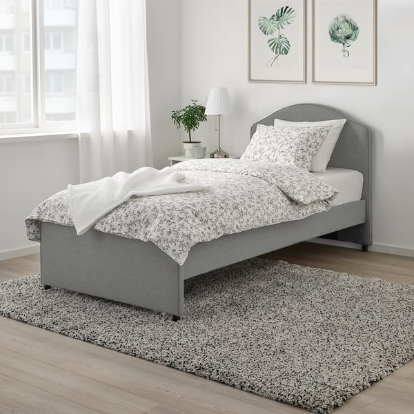 Graue Blumenbettwäsche auf gepflegtem Bett, graues Kopfteil, Teppich, Pflanzenkunst, Beistelltisch mit Lampe.