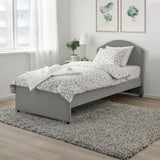 Graue Blumenbettwäsche auf gepflegtem Bett, graues Kopfteil, Teppich, Pflanzenkunst, Beistelltisch mit Lampe.