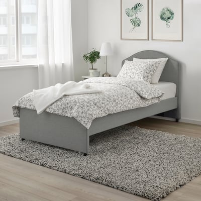 Graue Blumenbettwäsche auf gepflegtem Bett, graues Kopfteil, Teppich, Pflanzenkunst, Beistelltisch mit Lampe.