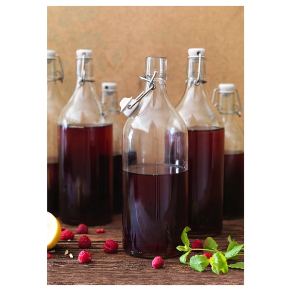 Klarglasflaschen mit Bügelverschluss, die dunkle Flüssigkeit enthalten, stehen auf einem Holztisch, auf dem Himbeeren und Minzblätter verstreut sind.