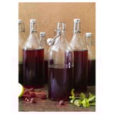 Klarglasflaschen mit Bügelverschluss, die dunkle Flüssigkeit enthalten, stehen auf einem Holztisch, auf dem Himbeeren und Minzblätter verstreut sind.
