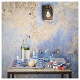 Klarsichtige KORKEN Glasflasche mit Zitrone und Gläsern. Verwitterte blaue Wand und Glühbirne. Gemusterte Textilien auf dem Tisch.