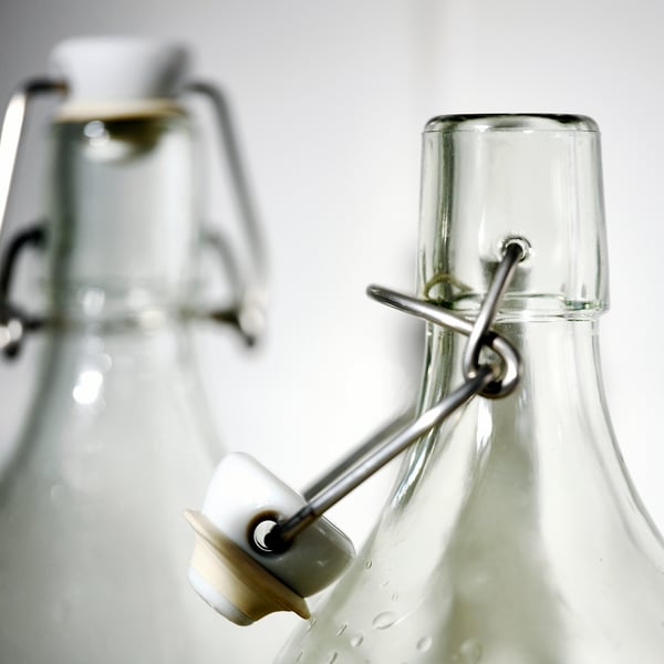 Klare Glasflaschen mit sicheren Metallklemmen und festen Stopfen. Eine Flasche scharf, eine andere im Hintergrund verschwommen.