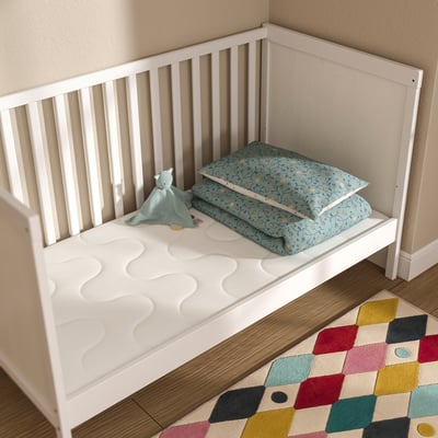 Weiße KRUMMELUR Krippenmatratze an der Wand, fest mit glatten Oberflächen, Stoff mit Reißverschluss, einfaches weißes Kinderbett, blaue Decke, Spielzeug, bunter Teppich.