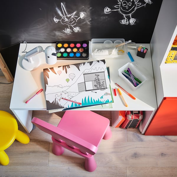 Basteltisch für Kinder mit KRUX-Lampe, farbigen Farben, Markern und rosa Stühlen.