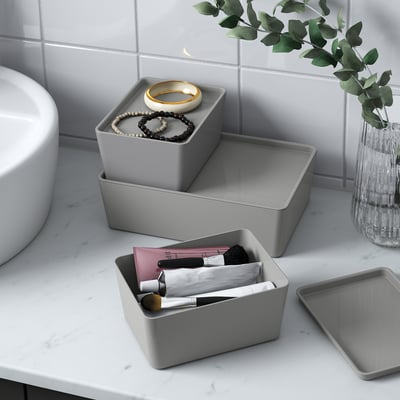 Drei graue KUGGIS-Boxen auf einer Badezimmertheke. Einer hält Make-up, ein anderer Schmuck und einer ist leer. Eine Vase mit Grün ist ebenfalls vorhanden.