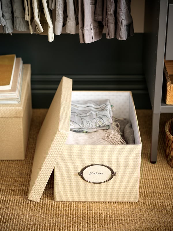 Eine beige KVARNVIK-Aufbewahrungsbox mit einem runden Etikett schals auf einem schwarzen Schrankboden, in dem gefaltete Schals aufbewahrt werden.
