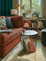 Helles Wohnzimmer mit braunem KVISTBRO-Beistelltisch mit Büchern, rotbraunem Sofa, blauen Vorhängen, Grün und Bücherregalen.