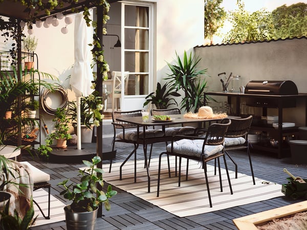 Gemütliche Terrasse LÄCKÖ schwarze Stühle mit grauen Kissen um einen Tisch, umgeben von Pflanzen und Lichterketten, die einen entspannenden Essbereich im Freien schaffen.
