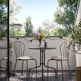 Moderner Balkon mit schwarzem Tisch, weißen Kissen, bunten Blumen und Lichterketten.
