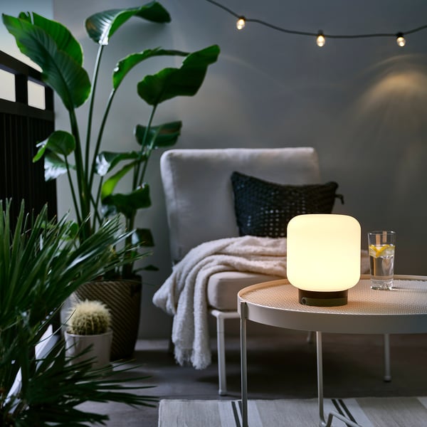 Gemütliches Wohnzimmer mit LÄNSPORT-Lampe auf Tisch, weißem Stuhl, Pflanzen, Lichterketten. Lampe klein im Vergleich zu anderen.