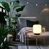 Gemütliches Wohnzimmer mit LÄNSPORT-Lampe auf Tisch, weißem Stuhl, Pflanzen, Lichterketten. Lampe klein im Vergleich zu anderen.
