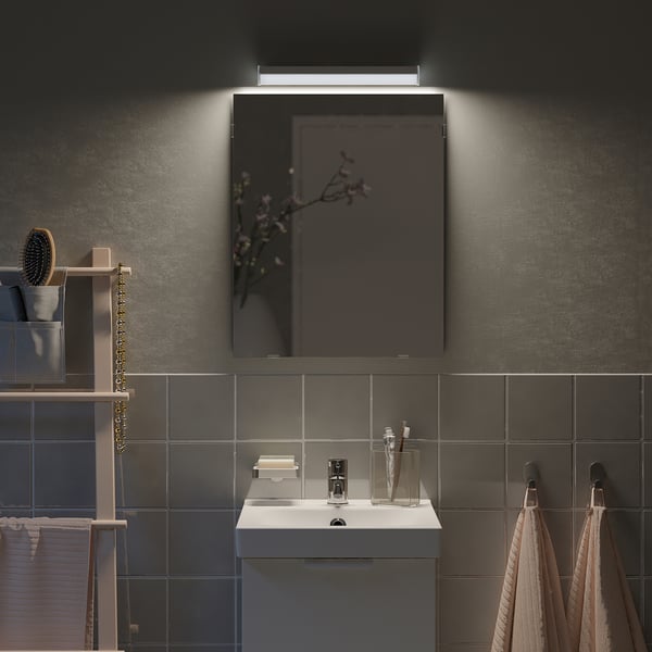 Kleiner, grauer LÄRBRO-Spiegel im Badezimmer, beleuchtet über weißem Waschbecken, umgeben von dunklen Fliesen.