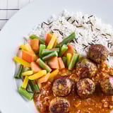 Teller mit pflanzlichen Fleischbällchen in Currysauce mit buntem gedünstetem Gemüse und Wildreis.