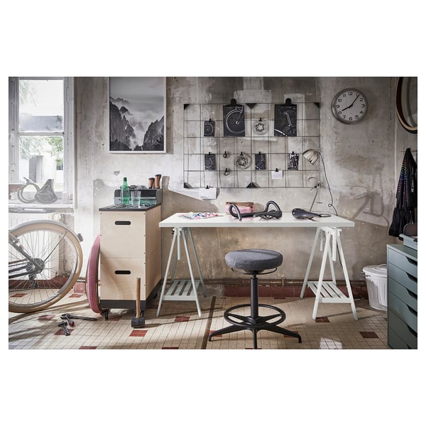 Arbeitsplatz im Industriestil mit weißem LAGKAPTEN-Schreibtisch, verstellbarem Hocker, Wanduhr und Dekor aus Fahrradteilen an einer Wand aus abgenutztem Beton.