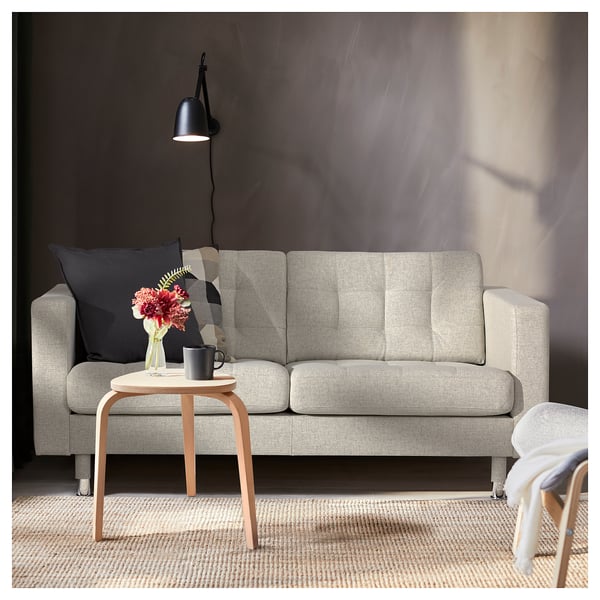 Modernes Wohnzimmer mit hellgrauem LANDSKRONA-Sofa, Beistelltisch aus Holz und schwarzer Wandleuchte. Dekorative Kissen und Vase mit Blumen auf dem Tisch.