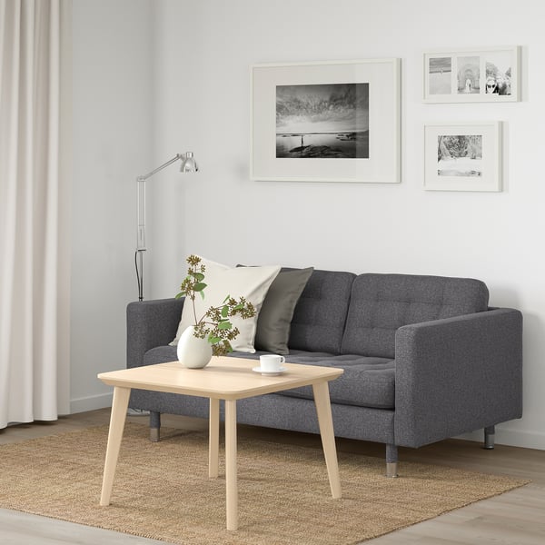 Gemütliches Wohnzimmer mit grauem Sofa, Holztisch, Lampe und Fotos. Weiße und schwarze Kissen, Blumen, leichte Vorhänge und beige Teppiche.