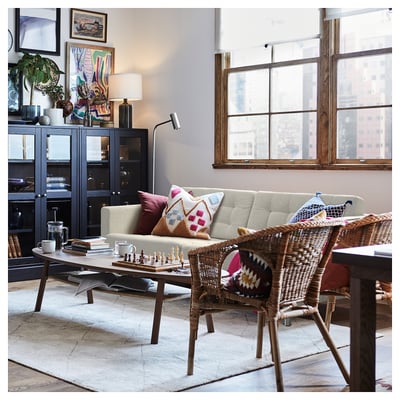 Modernes Wohnzimmer mit einem beigen LANDSKRONA-Sofa, einem geflochtenen Stuhl, einem Couchtisch aus Holz und großen Fenstern. Dekorative Kissen und ein Schachspiel auf dem Tisch.