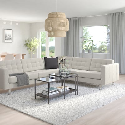Modernes Wohnzimmer mit Landnrona beigem Ecksofa, zwei Kissen, Couchtisch mit Glasplatte, großem Fenster und Pendelleuchte.