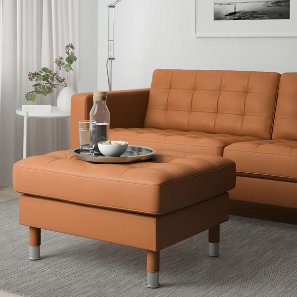 Orangefarbene lederähnliche Ottomane, passende große Couch, mit Getränketablett und Schüssel obendrauf, positioniert vor weißer Wand und Vorhang.