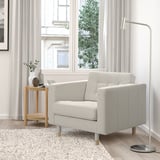 Modernes Wohnzimmer mit grauem LANDSKRONA-Sessel, Beistelltisch aus Holz, Stehleuchte und großem Fenster. Vorhänge und Teppiche vervollständigen das neutrale Dekor.