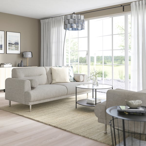 Modernes Wohnzimmer mit LÅNGARYD-Sofa, Glastisch, grauen Vorhängen, Lampe und Büchern. Große Fenster durchfluten es mit natürlichem Licht.