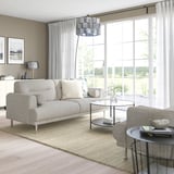 Modernes Wohnzimmer mit LÅNGARYD-Sofa, Glastisch, grauen Vorhängen, Lampe und Büchern. Große Fenster durchfluten es mit natürlichem Licht.