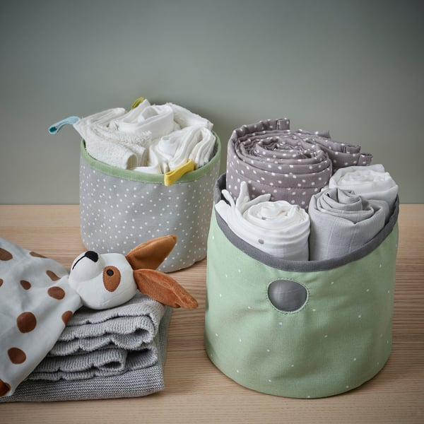 Zwei Stoffbehälter, ein grüner, ein grauer mit weißen Tupfen, in denen zusammengerollte Babykleidung und ein Stofftier auf einem Holztisch stehen.